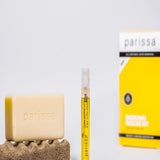 Pale rectangular Parissa bar resting on a textured brown pad beside a slim yellow-labelled tube, with the yellow Ingrown Rescue Kit box in the background.