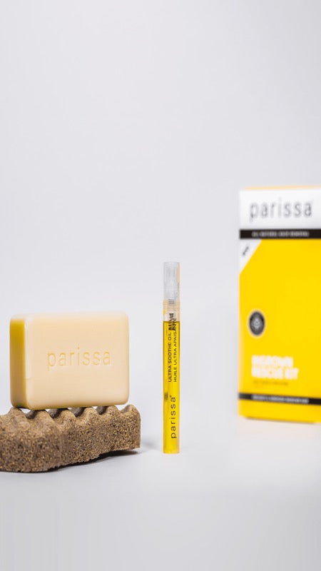 Pale rectangular Parissa bar resting on a textured brown pad beside a slim yellow-labelled tube, with the yellow Ingrown Rescue Kit box in the background.