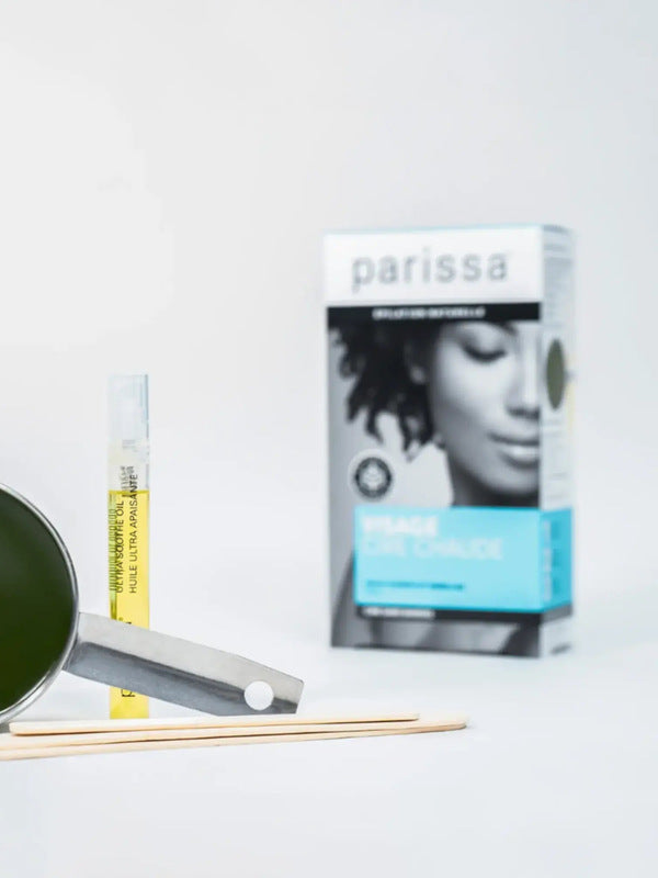 Small metal pan of dark green wax, wooden spatulas and a slim yellow-labelled tube arranged in front of a Parissa Face and Lip Hot Wax box.