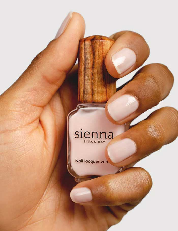 A hand with neatly manicured nails painted in Peace, a soft pale pink shade from Sienna Byron Bay Nail Polish (10ml), holds the bottle with its signature wooden cap.