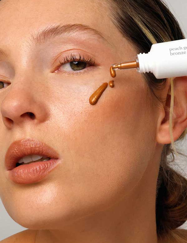 A woman applies Ere Perez Peach Gel Bronze Drops, a warm, golden brown shade, to her cheeks with a dropper. Her skin glows with natural makeup, nude lips, and soft eye makeup.