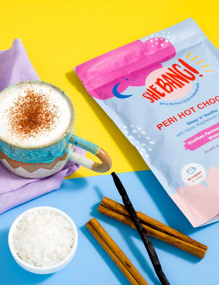 A vibrant scene shows She BANG! Peri Hot Choccy 300g for women, with a mug of hot chocolate topped with frothy milk and cinnamon, two cinnamon sticks, rock salt, and vanilla pods on a yellow and blue background for PMS support.