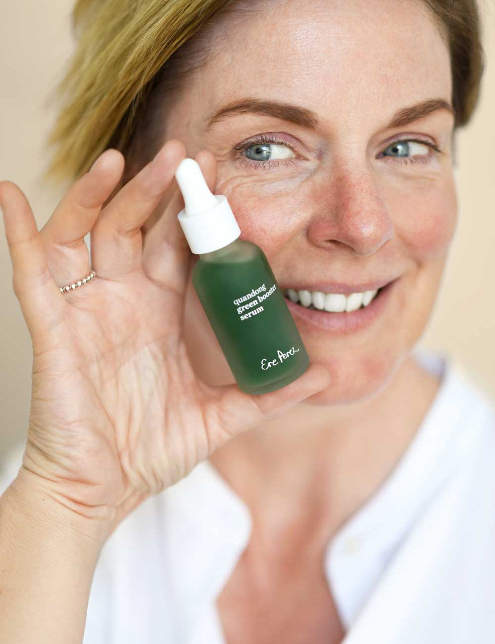 A smiling woman with short, light brown hair holds up Ere Perez Quandong Green Booster Serum 30ml, a vitamin C and hyaluronic acid antioxidant serum. She wears a white top and has a natural, minimal makeup look.