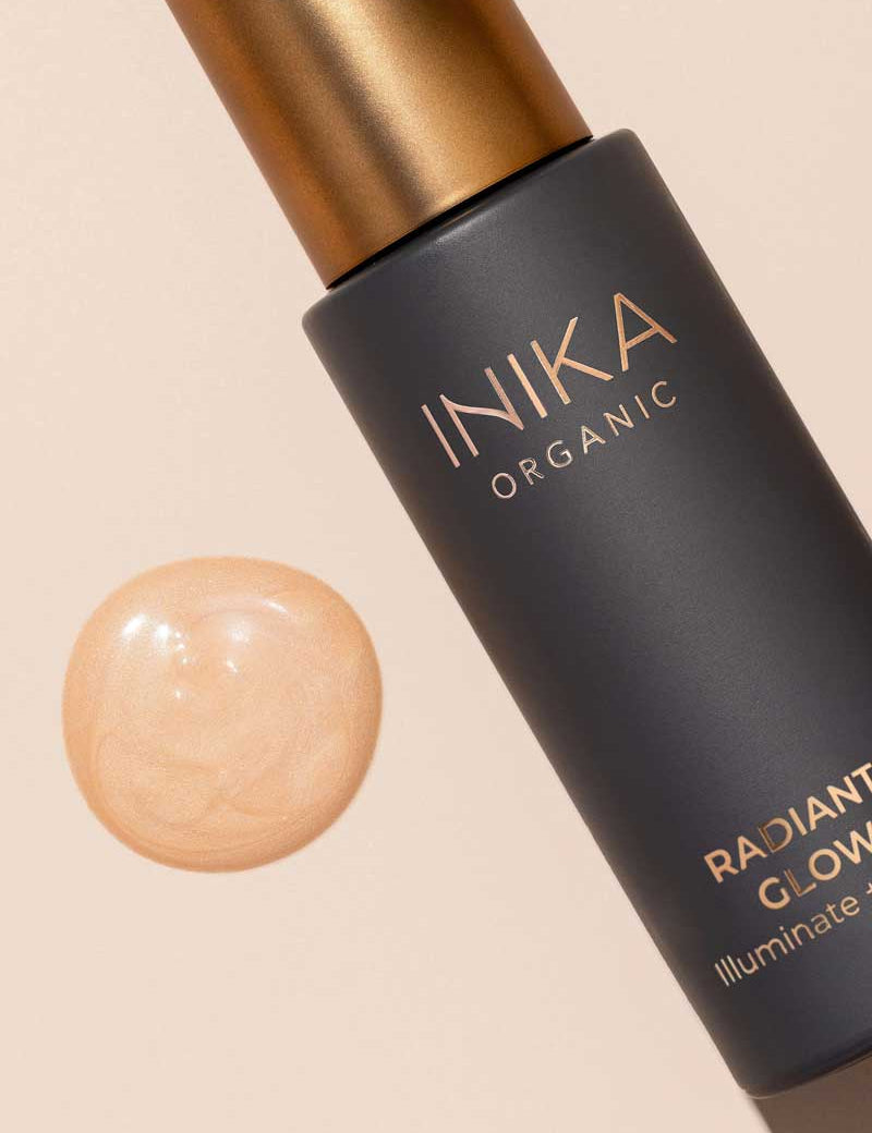 A bottle of INIKA Organic Radiant Glow Veil Primer 30ml rests beside a swatch of shimmery, light beige liquid on a neutral background.