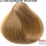 Rounded teardrop-shaped tuft of smooth, muted golden-beige hair beneath the shade name Champagne Blonde, with the Radico Colour Me Organic logo at the lower right.