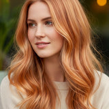 Woman outdoors with long, centre-parted peach-copper waves and pale golden highlights, with a small label beneath identifying the shade as Strawberry Blonde.