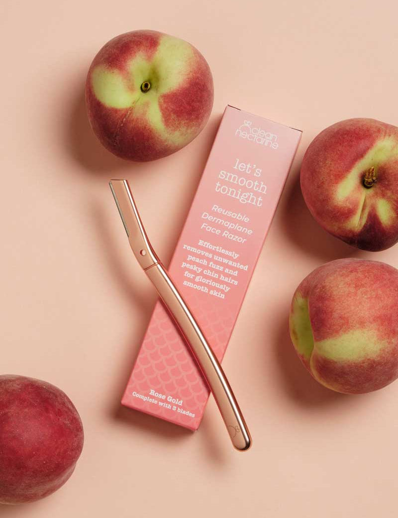 A Clean Nectarine Reusable Metal Dermaplaner in rose gold lies beside its pink packaging reading "let’s smooth tonight" and "Reusable Dermaplane Face Razor for skin exfoliation," surrounded by four ripe peaches on a beige background.