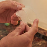 Hands peeling a translucent crescent-shaped under-eye patch away from a clear backing sheet.