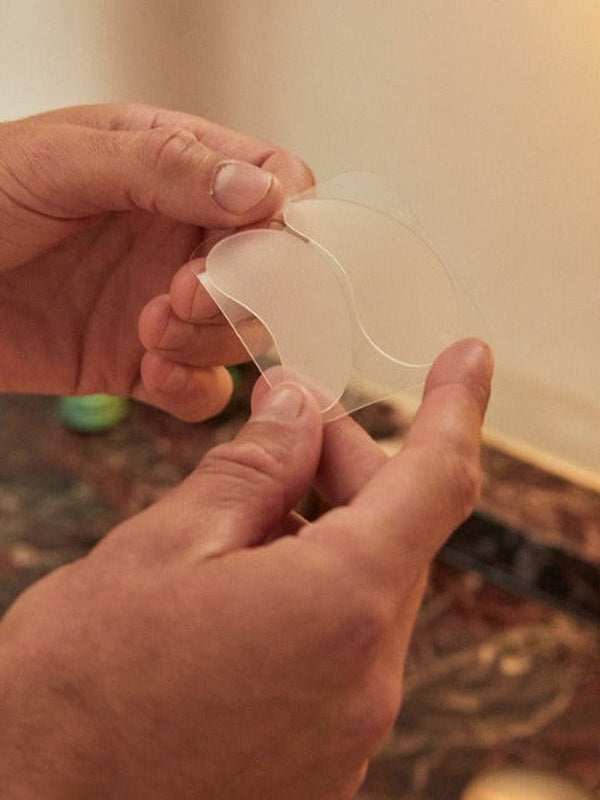 Hands peeling a translucent crescent-shaped under-eye patch away from a clear backing sheet.