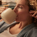 Woman reclining in a grey robe with translucent patches beneath her eyes, drinking from a large cream-coloured mug.