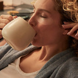 Woman reclining in a grey robe with translucent patches beneath her eyes, drinking from a large cream-coloured mug.