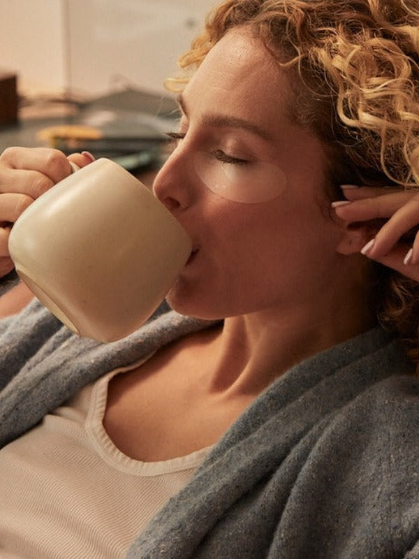 Woman reclining in a grey robe with translucent patches beneath her eyes, drinking from a large cream-coloured mug.