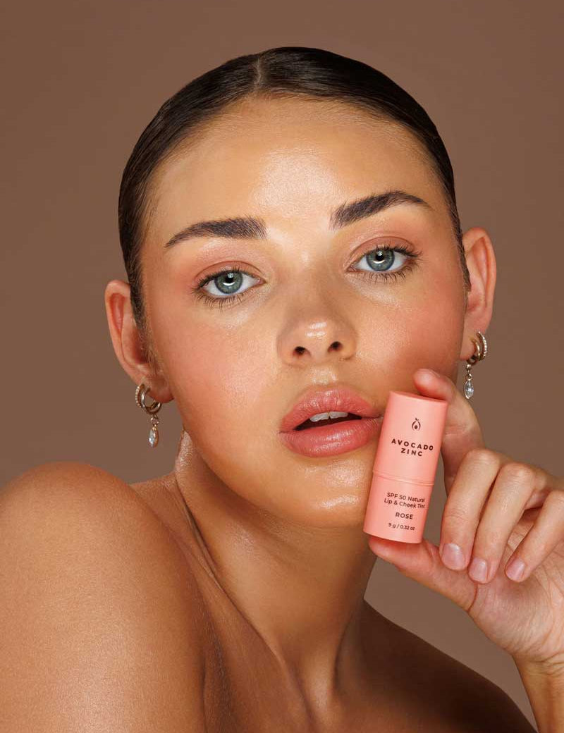 A woman with slicked-back hair holds the Avocado Zinc SPF50 Natural Lip & Cheek Tint, Rose 9g near her face. She has dewy makeup, glossy lips, and wears small hoop earrings against a brown backdrop.