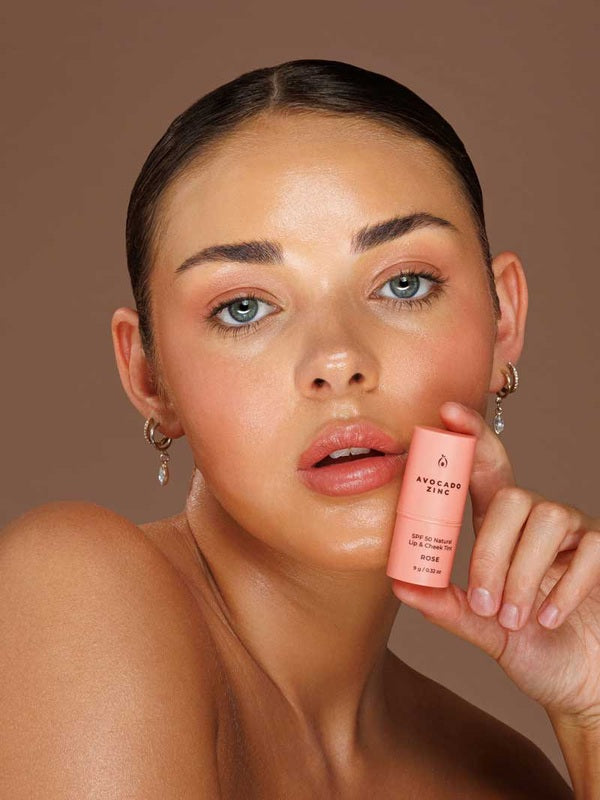 Model wearing Avocado Zinc SPF50 Natural Lip & Cheek Tint in Rose and holding the tint beside her face.
