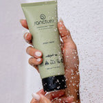 Sanctum Body Buff tube held in hands beneath running shower water during use.