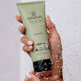 Sanctum Body Buff tube held in hands beneath running shower water during use.