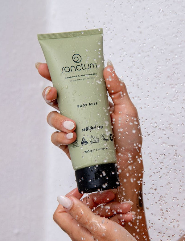 Sanctum Body Buff tube held in hands beneath running shower water during use.