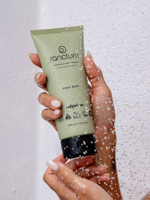 Sanctum Body Buff tube held in hands beneath running shower water during use.