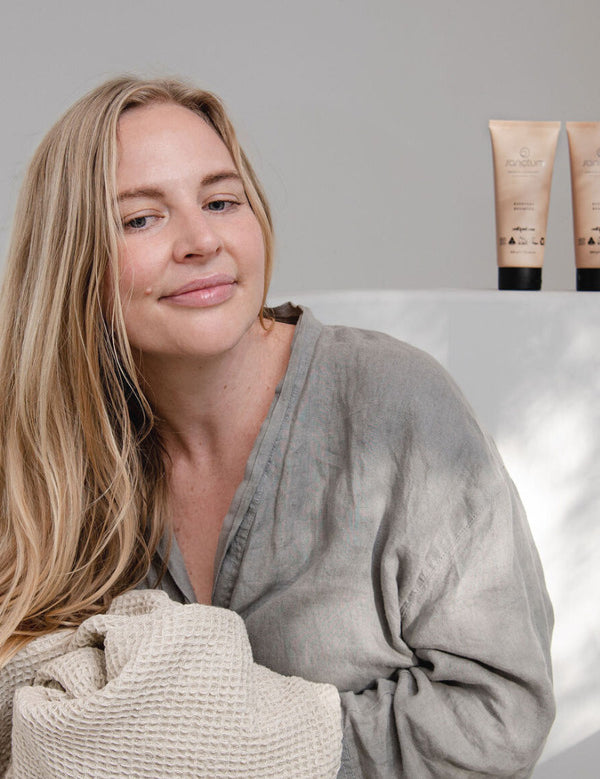 Woman in a grey robe holding a textured towel beside Sanctum Everyday Conditioner and matching haircare tubes.