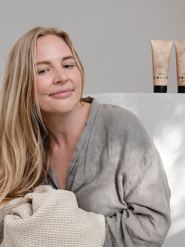 Woman in a grey robe holding a textured towel beside Sanctum Everyday Conditioner and matching haircare tubes.
