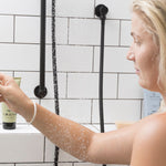 Woman using Sanctum Everyday Shampoo in a tiled shower, with Sanctum haircare tubes on the ledge.