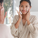 Woman gently exfoliating her face with Sanctum Gentle Face Exfoliant at a bathroom mirror.