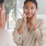 Woman gently exfoliating her face with Sanctum Gentle Face Exfoliant at a bathroom mirror.