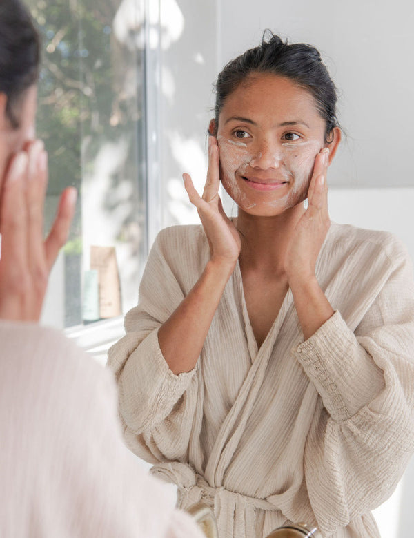 Woman gently exfoliating her face with Sanctum Gentle Face Exfoliant at a bathroom mirror.