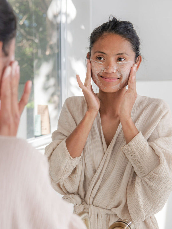 Woman gently exfoliating her face with Sanctum Gentle Face Exfoliant at a bathroom mirror.