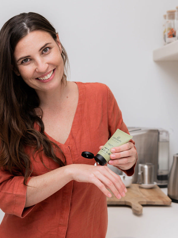 Woman applying Sanctum Hand and Nail Repair cream to her hands in a bright home setting.