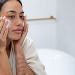 Woman applying Sanctum HY C Intensive Repair Cream to her cheek in front of a bathroom mirror.