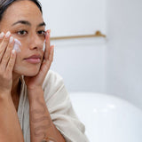 Woman applying Sanctum HY C Intensive Repair Cream to her cheek in front of a bathroom mirror.