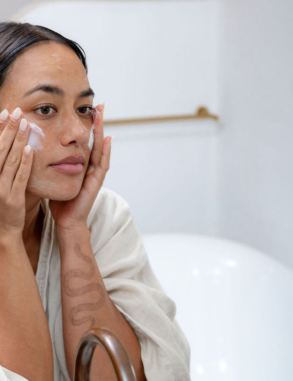 Woman applying Sanctum HY C Intensive Repair Cream to her cheek in front of a bathroom mirror.