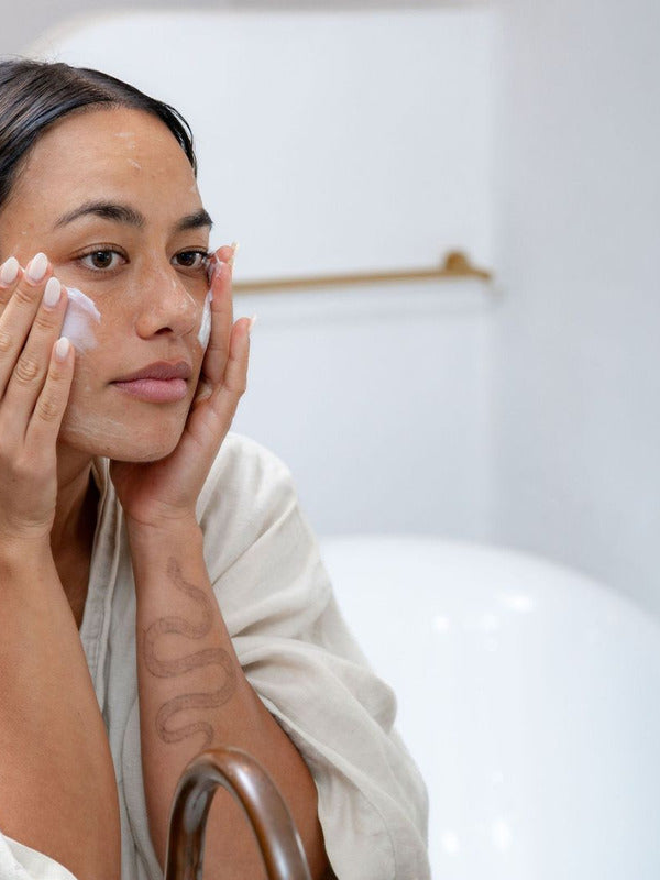 Woman applying Sanctum HY C Intensive Repair Cream to her cheek in front of a bathroom mirror.