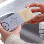 Hands removing a pale Sanctum Lavender and Chamomile Soap 100g bar from its cardboard box beside a kitchen sink.