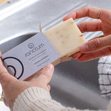 Hands removing a pale Sanctum Lavender and Chamomile Soap 100g bar from its cardboard box beside a kitchen sink.