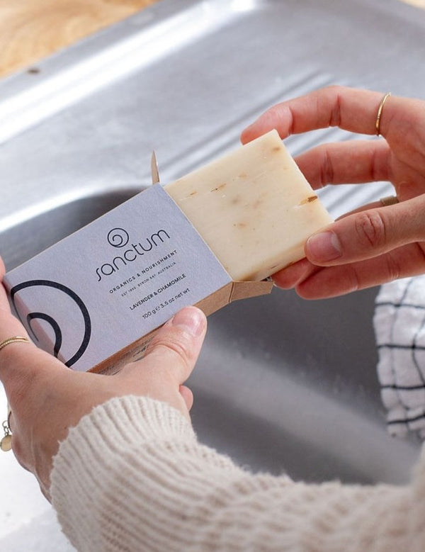 Hands removing a pale Sanctum Lavender and Chamomile Soap 100g bar from its cardboard box beside a kitchen sink.