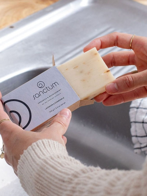 Hands removing a pale Sanctum Lavender and Chamomile Soap 100g bar from its cardboard box beside a kitchen sink.