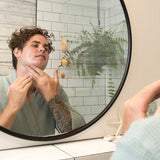 Man applying Sanctum Men's Face Moisturiser to his cheek while looking into a round bathroom mirror.