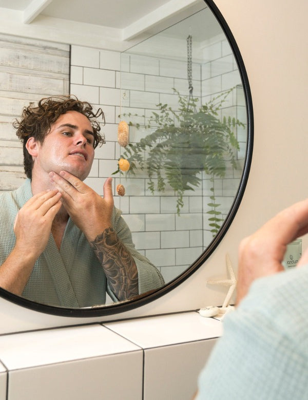 Man applying Sanctum Men's Face Moisturiser to his cheek while looking into a round bathroom mirror.