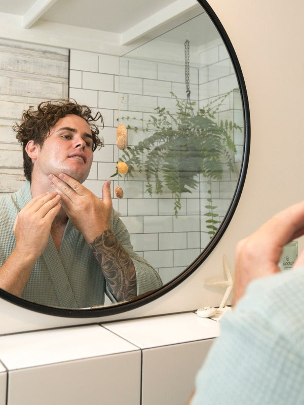 Man applying Sanctum Men's Face Moisturiser to his cheek while looking into a round bathroom mirror.