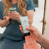 Woman dispensing Sanctum Moisture Balance moisturiser from a pale tube into her open palm in soft natural light.