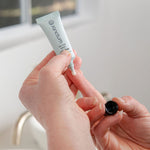 Hands holding the Sanctum On The Spot treatment tube and applicator beside a bathroom sink in soft natural light.