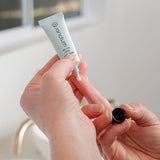 Hands holding the Sanctum On The Spot treatment tube and applicator beside a bathroom sink in soft natural light.