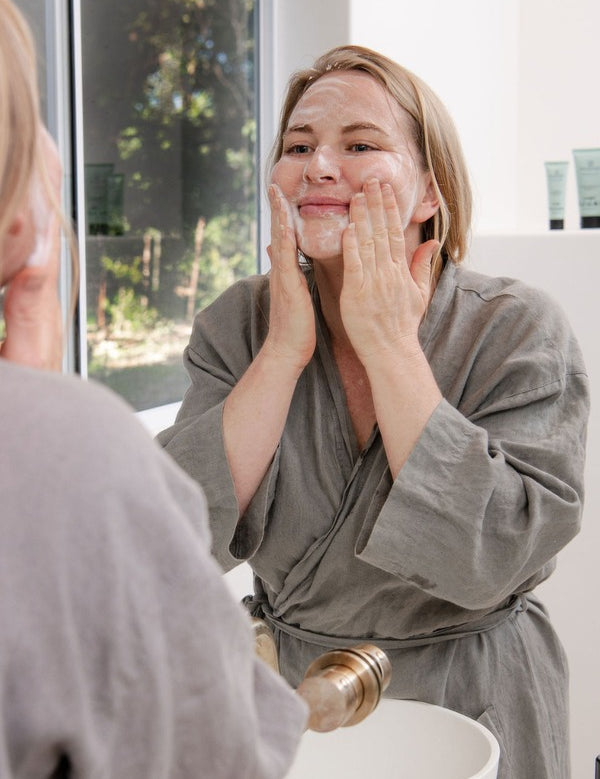 Woman in a grey robe cleansing her face with Sanctum Purifying Foam Cleanser while looking into a bathroom mirror.