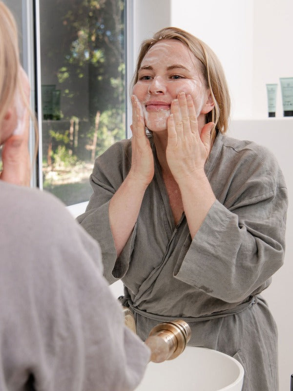 Woman in a grey robe cleansing her face with Sanctum Purifying Foam Cleanser while looking into a bathroom mirror.