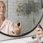 Woman spraying Sanctum Tone & Refresh Mist toward her face while looking into a bathroom mirror.