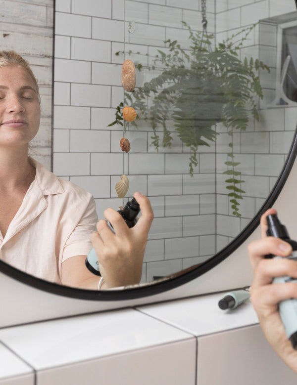 Woman spraying Sanctum Tone & Refresh Mist toward her face while looking into a bathroom mirror.