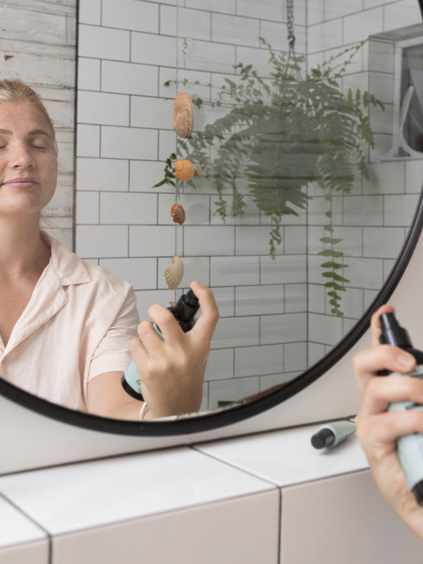 Woman spraying Sanctum Tone & Refresh Mist toward her face while looking into a bathroom mirror.