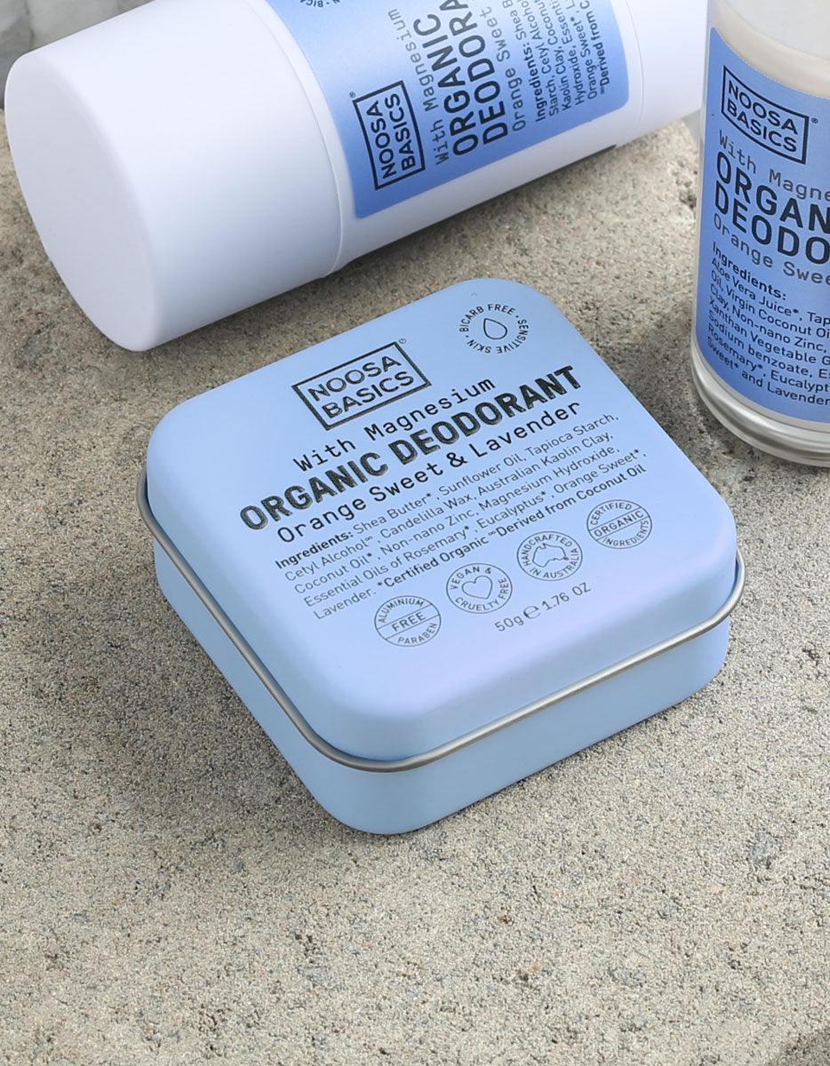Close-up of pale blue Noosa Basics Sweet Orange and Lavender deodorants on a stone surface, with a square tin in front of a white stick container and a roll-on bottle.